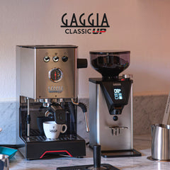 Stainless steel Gaggia Classic UP espresso machine beside a Gaggia coffee grinder, with a cup, tamper and milk jug.