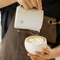 Barista pouring latte art into white cup with white Timemore Rosetta Pro Milk Pitcher