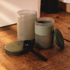 Sage Green Wacaco Prestina with a cup of coffee, caps and scoop on a wooden table.