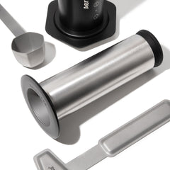 AeroPress stainless steel plunger, coffee scoop and stirrer beside the black brewing chamber.