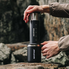 Person pressing an AeroPress Stainless Steel Coffee Press into a black mug on a rock outdoors.