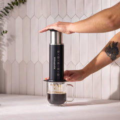 Person pressing an AeroPress Stainless Steel Coffee Press into a glass mug on a kitchen counter.