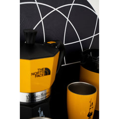 Close-up of the yellow and black Bialetti x The North Face moka pot beside a yellow cup and coffee tin.