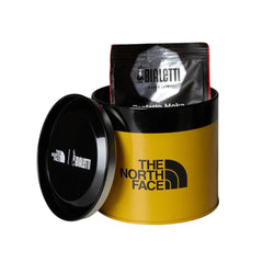 Open yellow The North Face coffee tin holding a Bialetti coffee bag, with the black co-branded lid alongside.