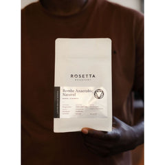 Person holding a white bag of Rosetta Roastery Bombe Anaerobic Natural coffee against a brown shirt.
