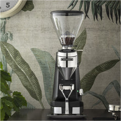Black Mazzer Super Jolly g espresso grinder on a café counter against a leafy patterned wall.