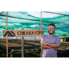 Tadiru Tedesse standing beside a Chilaka Coffee sign under green shade netting at the drying beds