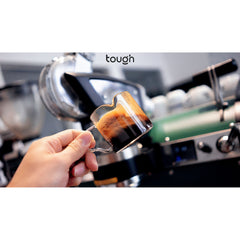 Hand swirling coffee inside Tough Double Spout Espresso shot glass with coffee machine in background