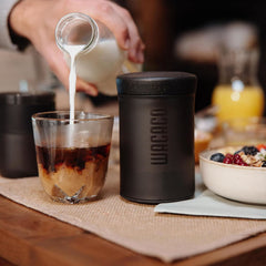 Pouring milk into a glass of coffee beside the packed Wacaco Prestina and a breakfast bowl.