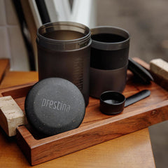 Wacaco Prestina brewing chamber, cup, cap and scoop separated on a wooden tray.
